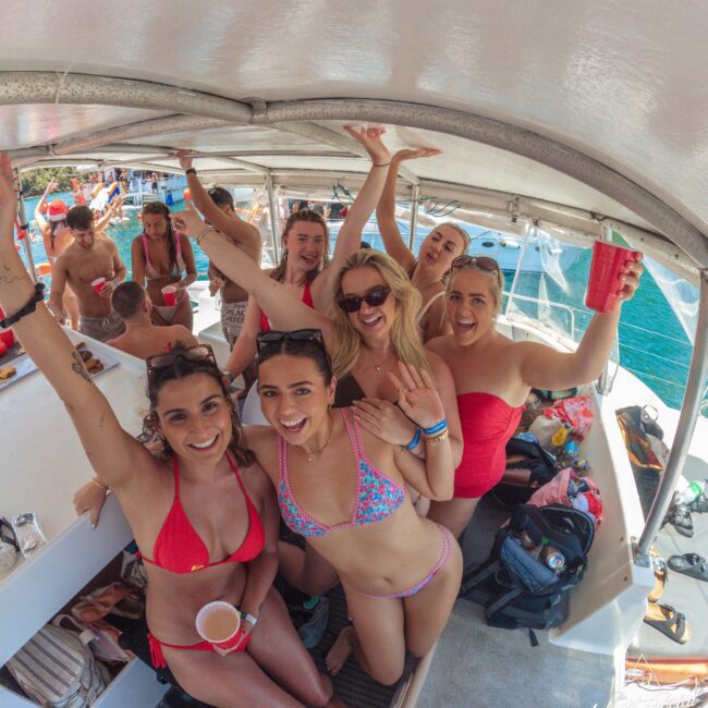 A group of smiling young women in swimsuits pose together on a boat, holding drinks and raising their arms. Other people relax in the background, with clear blue water and boats visible outside. The scene is lively and festive.