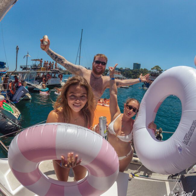 Three people in swimsuits hold inflatable rings and drinks, smiling on a boat with others and boats in the background, enjoying a sunny day on the water.