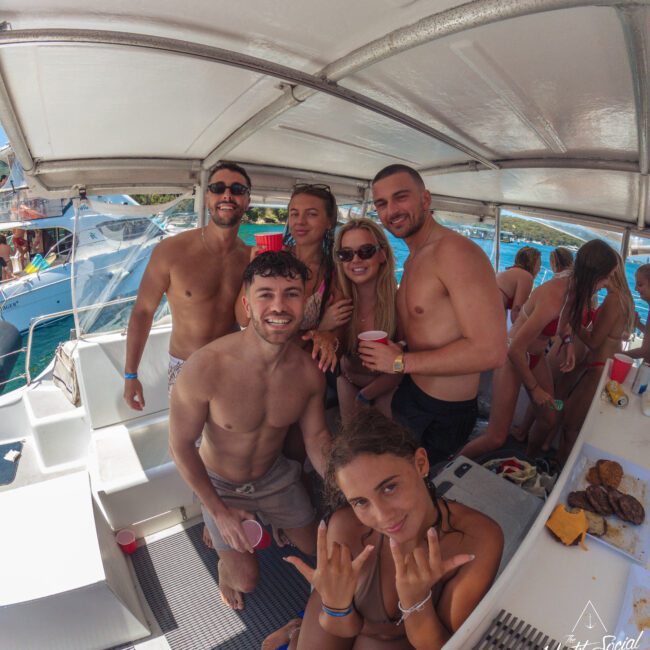 A group of young adults in swimsuits pose and smile on a boat, with tropical water and other boats in the background. Some are holding drinks, and one person sits in front making a hand gesture. It is sunny and lively.