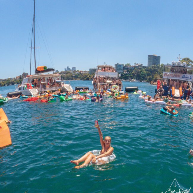 A woman in sunglasses floats on an inner tube in a blue body of water, holding up a drink. Behind her, people relax on inflatables and socialize near anchored yachts, with city buildings and trees in the background.