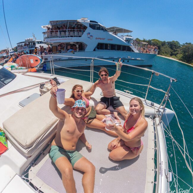Four young adults in swimwear sit on the deck of a yacht, smiling and raising drinks. Behind them, other boats and people are visible on the water, suggesting a lively, sunny boating event or party.
