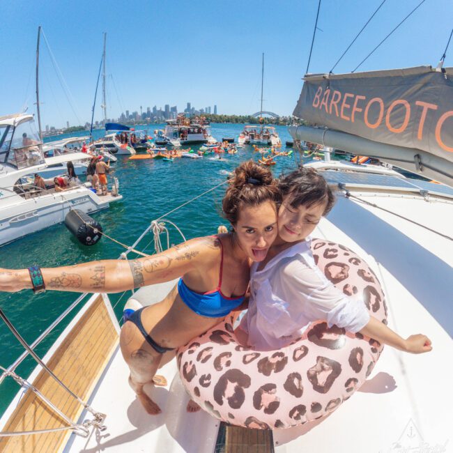 Two women smiling on a boat; one in a blue bikini, the other in a white shirt inside a pink leopard-print float. The boat is surrounded by water and other boats, with a city skyline in the background.