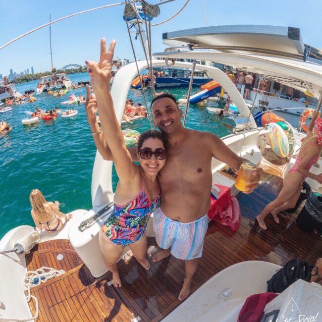 A smiling woman in a colorful swimsuit and a man in swim trunks stand on a boat, posing for a photo at a lively boat party with people swimming and relaxing on the water in the background.