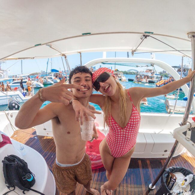 Two people in swimwear smile and pose for the camera on a boat, surrounded by other boats and people on the water. The scene is lively and summery, with blue skies and a festive atmosphere.