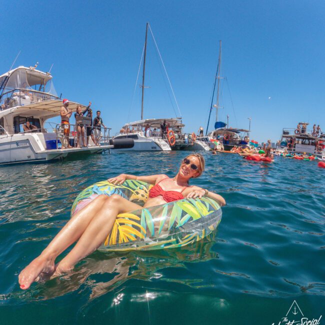 A woman in sunglasses and a red swimsuit relaxes on a tropical-patterned float in the water, surrounded by anchored yachts and other people enjoying a sunny day at a boat party.