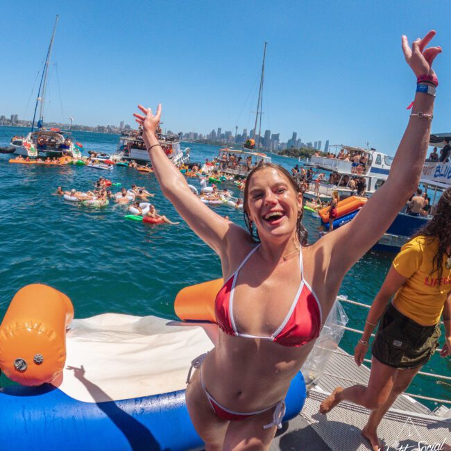 A smiling woman in a red bikini poses with arms raised on a boat, surrounded by people on inflatables and other boats on a sunny day, with a city skyline in the background.