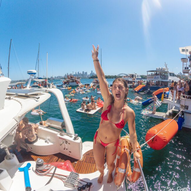 A woman in a red bikini stands on a boat, smiling and flashing a peace sign. Behind her, many people enjoy a sunny day on boats and inflatables in crowded blue water, with a city skyline visible in the distance.