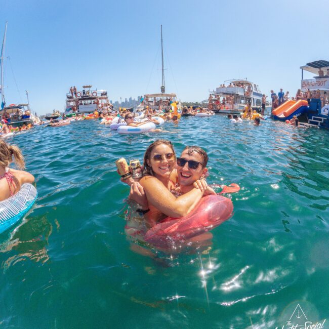 A smiling couple hugs while floating on pool inflatables in a lively gathering of people swimming around boats on a sunny day. Other boats and swimmers are in the background under a clear blue sky.