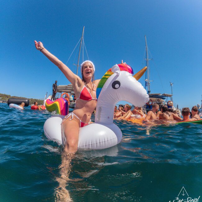 A woman in a red bikini smiles and raises her arm while sitting on a unicorn float in the ocean. People on other inflatables and boats are in the background under a clear blue sky.
