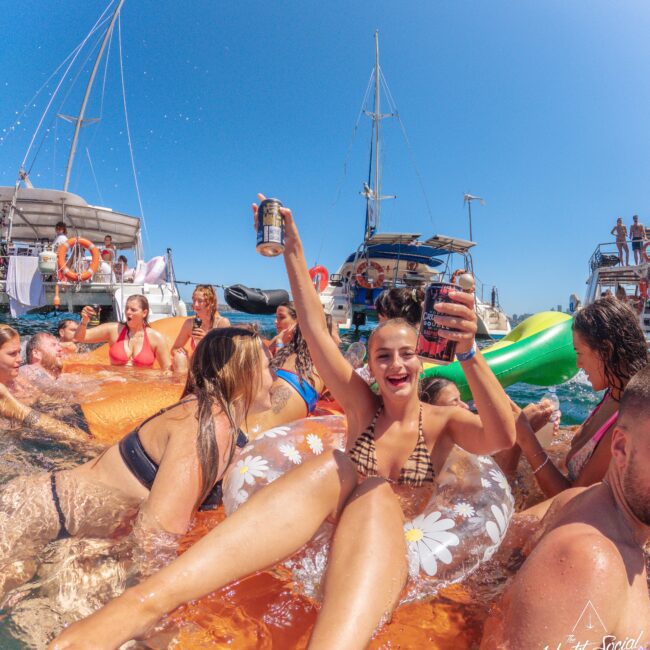 A group of people in swimsuits enjoy a lively party in the water near boats. One woman in a floral float holds up two drinks, smiling and celebrating, surrounded by others laughing and swimming under a clear blue sky.