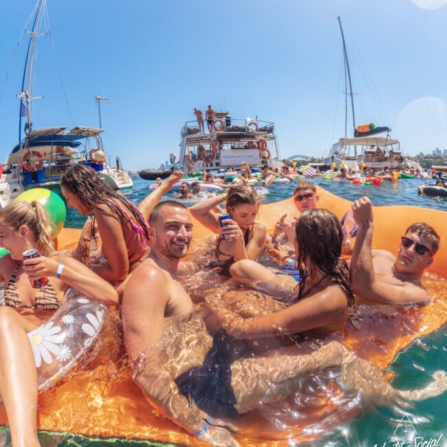 A group of people relax and socialize on an orange floating mat in the water near several docked yachts on a sunny day, holding drinks and enjoying a lively atmosphere.