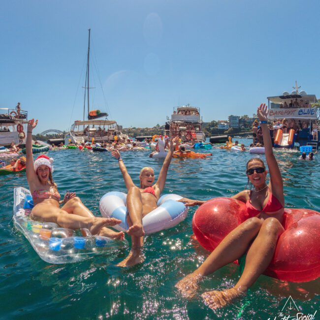 Three women in swimsuits float on colorful inflatables, smiling and waving, surrounded by boats and other people enjoying a sunny day on the water. The scene is festive and lively under a clear blue sky.
