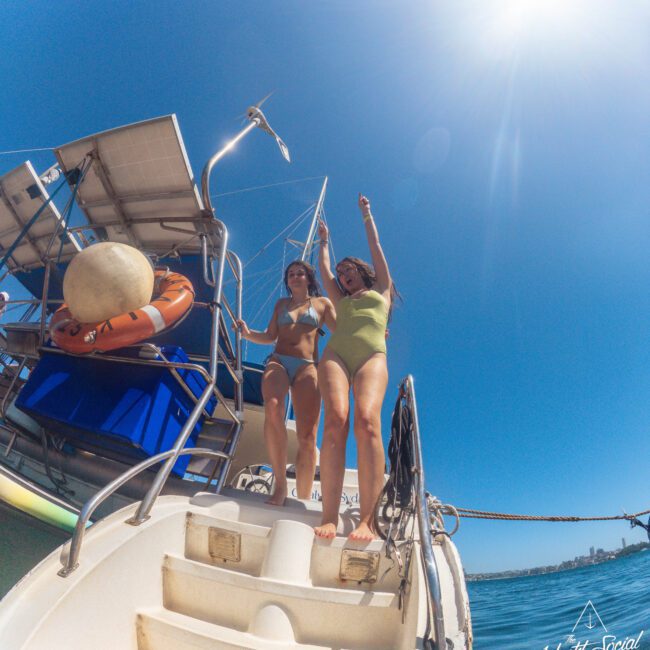 Two women in swimsuits stand on the back steps of a yacht under a bright blue sky, smiling and pointing excitedly. The sun is shining, and the sea and distant coastline are visible in the background.