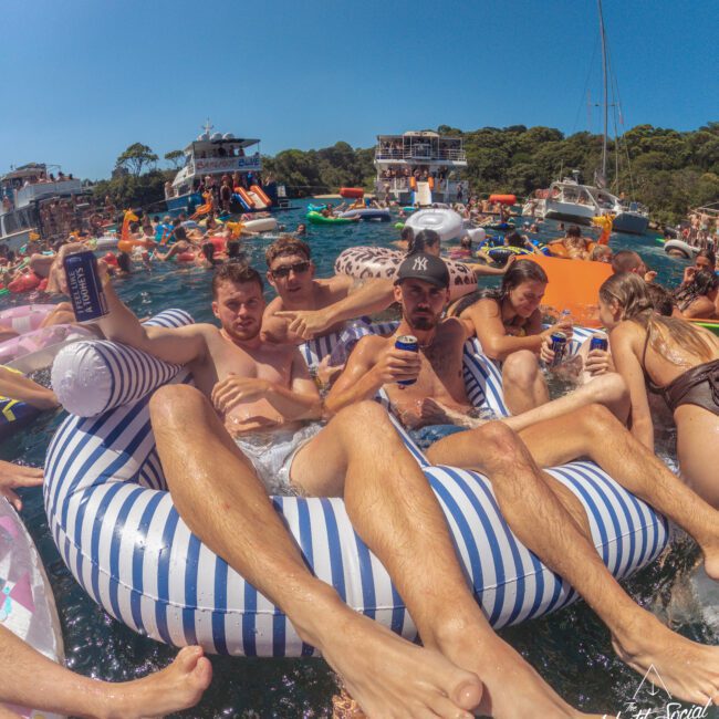 A group of young adults lounge on a large striped inflatable in a crowded, lively boat party on the water, surrounded by other floaties, boats, and people under a clear blue sky.