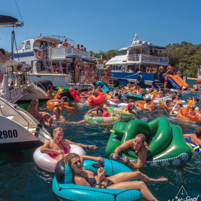 A lively scene of people relaxing on colorful inflatables in the water near several docked boats, enjoying sunny weather at a boat party. The background shows trees and clear blue sky.