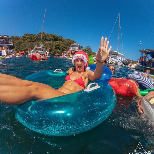 A woman in a red bikini and Santa hat smiles and waves while lounging in a blue inflatable ring on the water, surrounded by other people on floats and boats, with clear blue sky above.
