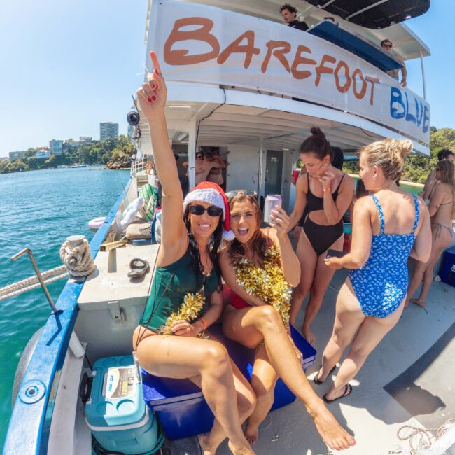 Two women in swimsuits sit on a boat, smiling and celebrating with gold tinsel, while others in swimsuits chat nearby. The boat reads "Barefoot Blue," and the scene is sunny with blue water and buildings in the background.