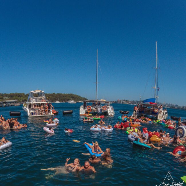 A large group of people enjoy a sunny day in the water on colorful inflatables near anchored boats, with green hills in the background and clear blue skies overhead. A "Yacht Social Club" logo appears at the bottom right.