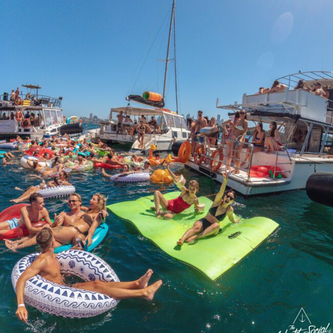A lively crowd enjoys a sunny day on the water, lounging on colorful floaties and rafts near anchored boats. People are laughing, relaxing, and socializing at a vibrant boat party.