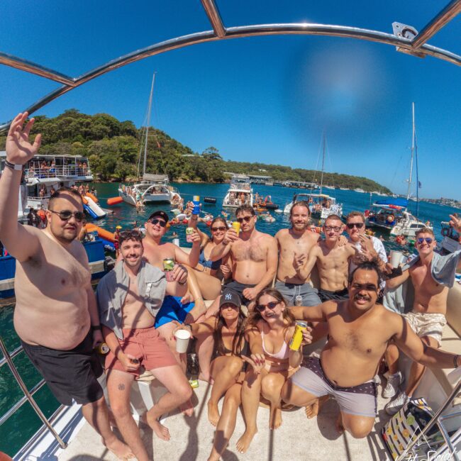 A group of people in swimsuits smile and pose on a boat under a sunny blue sky, surrounded by other boats and people on the water, with green trees in the background.