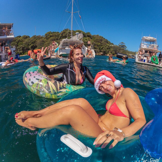 Two women relax on inflatable pool floats in the ocean near yachts. One wears a Santa hat and red bikini, smiling at the camera, while the other waves. Other people enjoy the water and boats in the background.