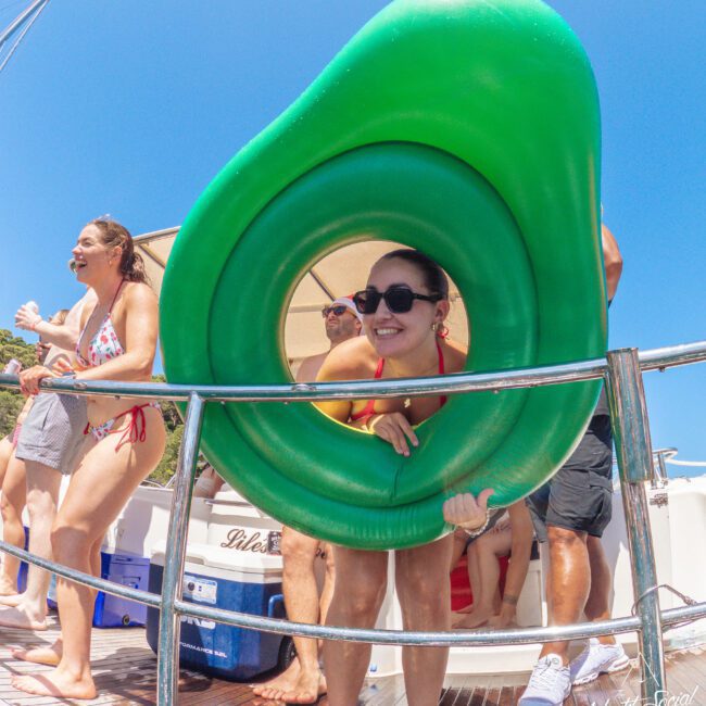A smiling woman wearing sunglasses leans through a large inflatable avocado pool float on a boat, with other people in swimwear enjoying the sunny day around her.