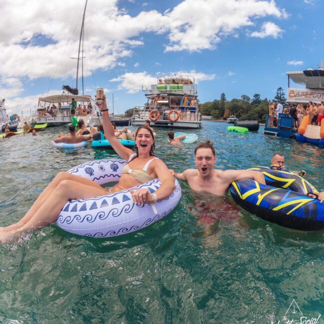 A smiling woman and man relax on inflatable tubes in the water, holding drinks. Other people and boats are visible in the background under a partly cloudy sky. The scene is lively and festive.