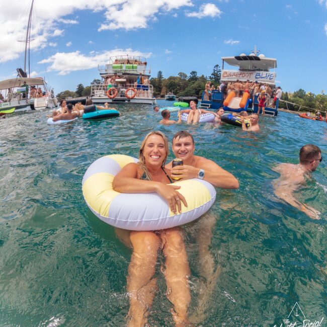 A smiling couple relaxes in a yellow and white pool float in clear blue water, surrounded by people swimming and floating near several boats under a partly cloudy sky.