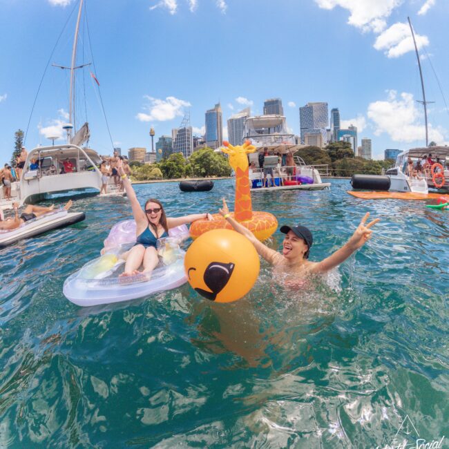 Two women enjoy a sunny day on the water; one on an inflatable float and the other holding an inflatable toy, surrounded by boats and a city skyline in the background.