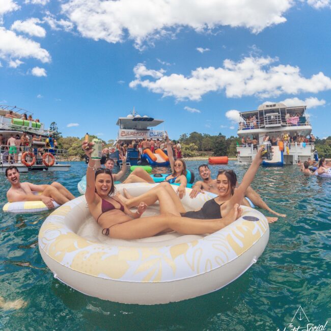 A group of young adults relax on a large inflatable float in the water, smiling and raising drinks, with boats and more people in the background under a bright, sunny sky.