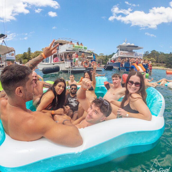 A group of young adults relax and smile on a blue and white inflatable floating in the water, with boats, people, and a bright sky in the background. Everyone appears to be enjoying a sunny, social day.