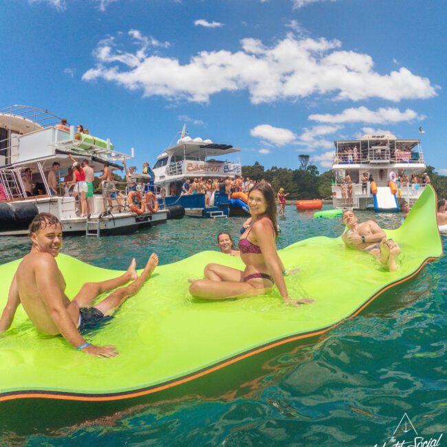 A group of young adults relax and smile on a large green floating mat in the water, surrounded by boats and people under a sunny blue sky. The scene appears lively and festive.