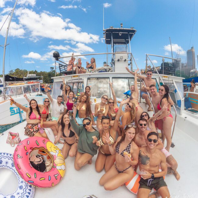A group of young adults in swimsuits smile and pose for a photo on a boat deck with pool floats and drinks, enjoying a sunny day. Some people stand on an upper deck, and city buildings are visible in the background.