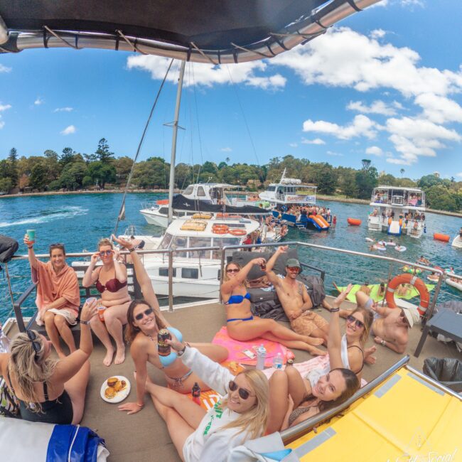 A group of people relax and smile on a boat, holding up drinks and enjoying snacks. Other boats and people swimming are visible in the water on a sunny day, with trees and a blue sky in the background.