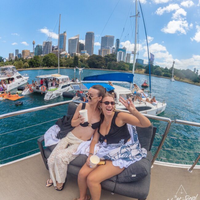 Two women sit on a yacht, smiling and taking a selfie while holding drinks. Other boats and people are in the water nearby, with tall city buildings and trees in the background under a sunny sky.