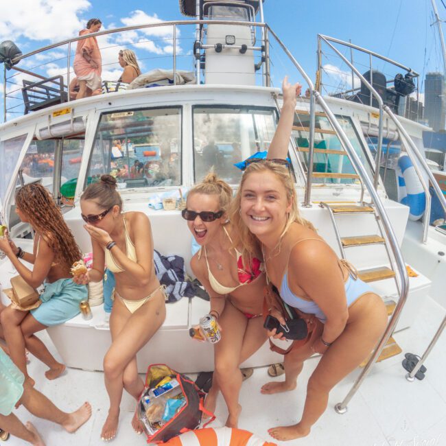 Three young women in swimsuits smile and pose on the deck of a boat, surrounded by friends and snacks, with blue skies and city buildings visible in the background.
