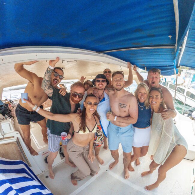A group of friends in swimsuits and summer clothes smile and pose together on a boat under a blue canopy, enjoying a sunny day on the water. Some have sunglasses and drinks. Other boats are visible in the background.