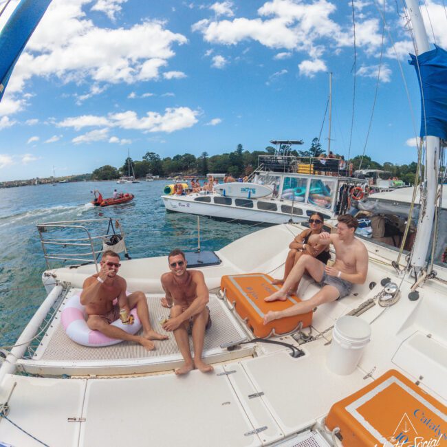 Five people in swimsuits relax on the deck of a catamaran yacht, surrounded by water and other boats, under a sunny blue sky with scattered clouds. Some are sitting on orange cushions and chatting together.