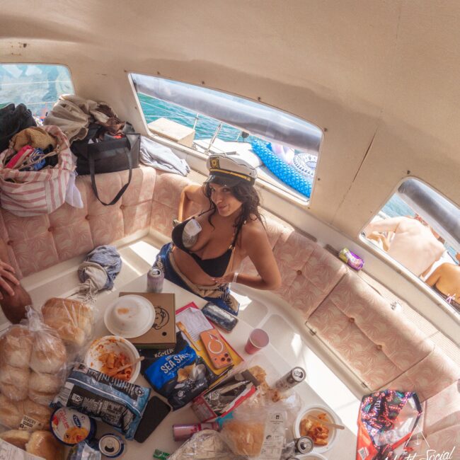 A woman in a captain’s hat and bikini poses and smiles inside a boat cabin, surrounded by snacks, drinks, and food on a cluttered table, with the sea visible through the windows.