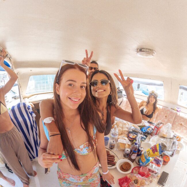 Two young women smiling and making peace signs pose on a boat with a table full of snacks and drinks. Other people are in the background, enjoying the gathering under bright daylight.
