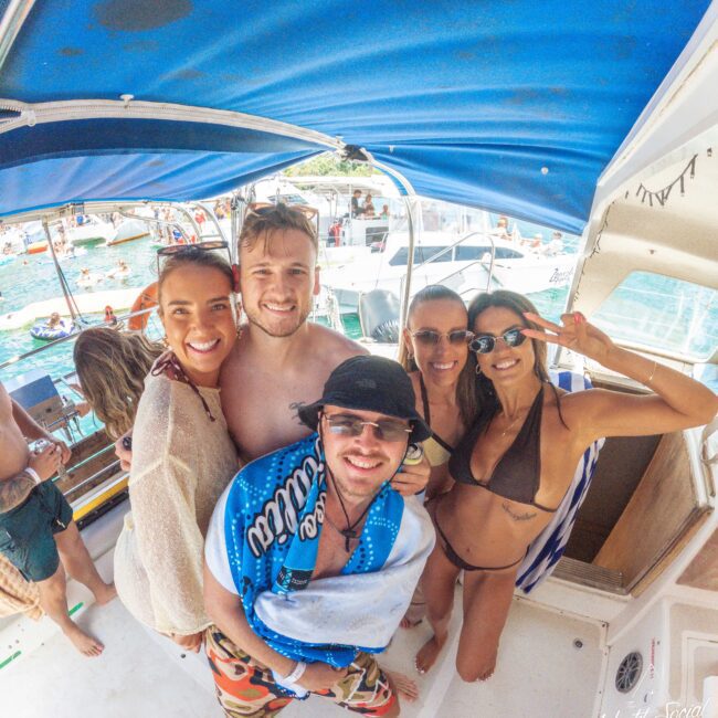 A group of five friends smiling and posing together on a boat under a blue canopy, with people and other boats visible on the water in the sunny background.