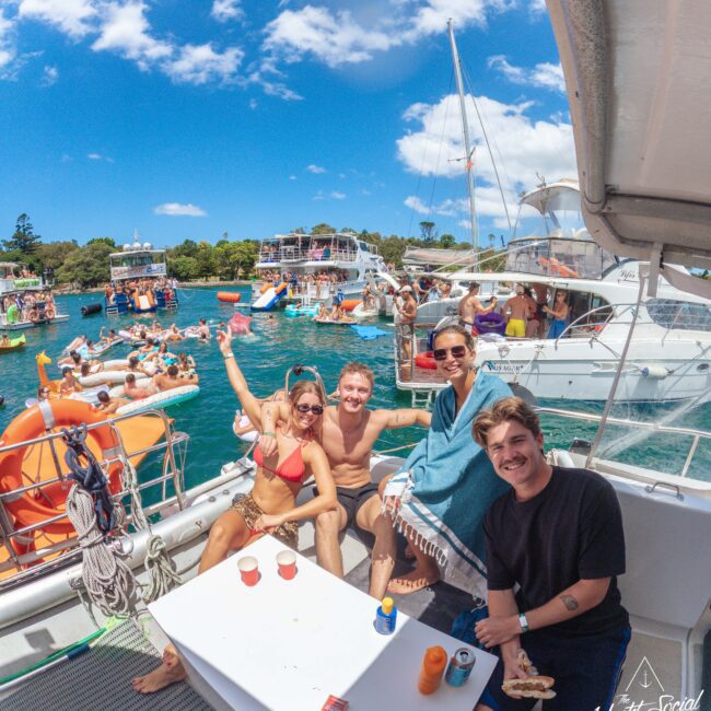 A group of smiling friends relax on a boat, enjoying food and drinks, with many other boats and people swimming and partying in the bright blue water under a sunny sky.
