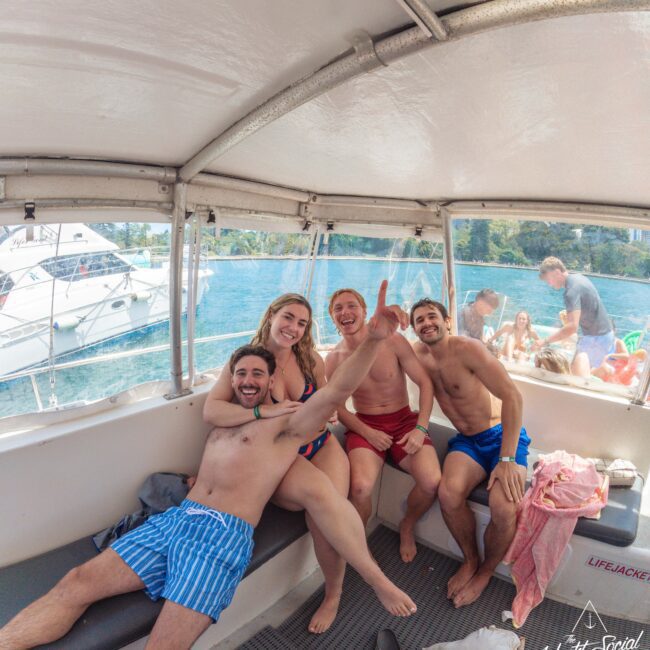 Four friends in swimwear smile and pose for a photo on a boat, with one person pointing at the camera. The boat is on blue water, with other boats and people visible in the background.