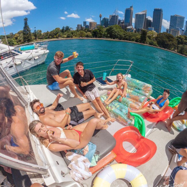 A group of young adults relax and smile on a yacht, some sitting and some lying on colorful pool floats. The city skyline and clear blue water are visible in the background under a sunny sky.