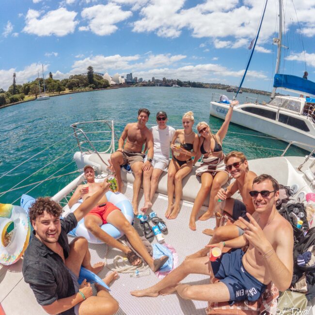 A group of young adults relax and smile on the deck of a yacht, enjoying drinks and the sunny weather, with blue water and city buildings visible in the background.