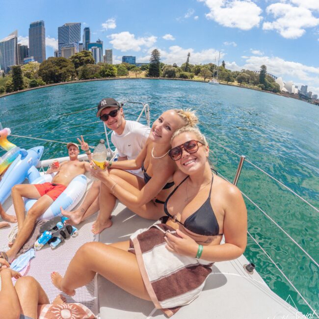 Four young adults relax on a boat in swimwear, smiling at the camera. City buildings and greenery are visible across the water on a sunny day with blue sky and clouds. Inflatable pool floats are beside them.