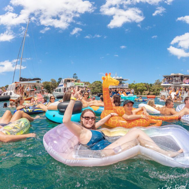 People relaxing on colorful inflatable floats in the water at a lively boat party under a sunny blue sky, with boats and more partygoers in the background. A smiling woman in sunglasses poses on a float in the foreground.