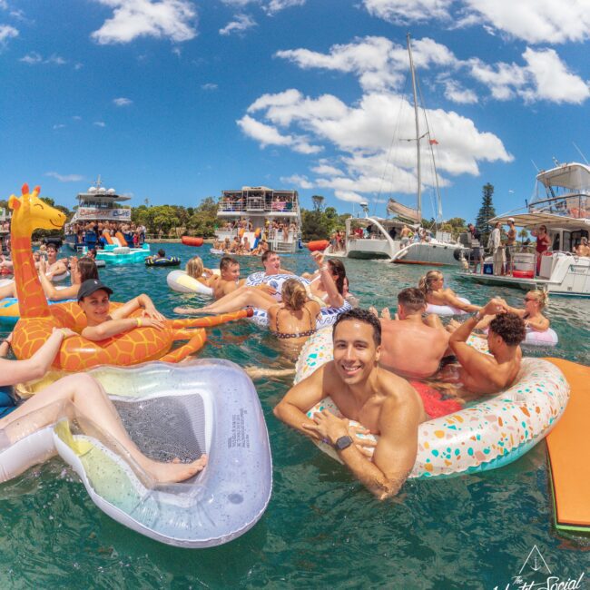 A group of people enjoy a sunny day on the water, floating on colorful inflatables near several anchored boats. The sky is blue with scattered clouds, and everyone appears relaxed and cheerful.