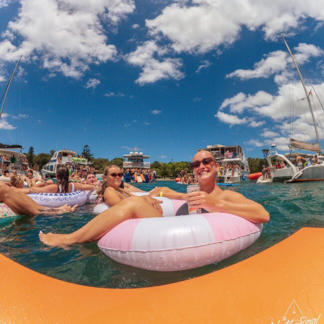 Two people relax on inflatable pool floats in the water, surrounded by boats and other people. The sky is blue with scattered clouds, and one person is smiling while holding a drink. “Yacht Social” is visible in the bottom right corner.