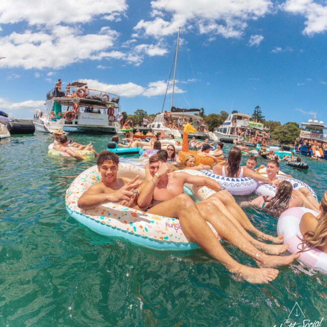 People relax on inflatables in the water near several anchored boats on a sunny day. The scene is lively, with groups enjoying the event, surrounded by clear blue water and a bright sky.